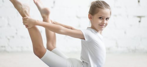 Smiling girl child practicing yoga, stretching in dhanurasana exercise, bow pose, working out wearing sportswear, t-shirt, pants, indoor full length, white loft studio background